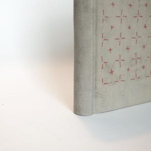 May include: A light gray book with a textured cover. The cover features a repeating pattern of red cross-like designs. The spine is rounded, and the book appears to be closed, with the pages unseen.