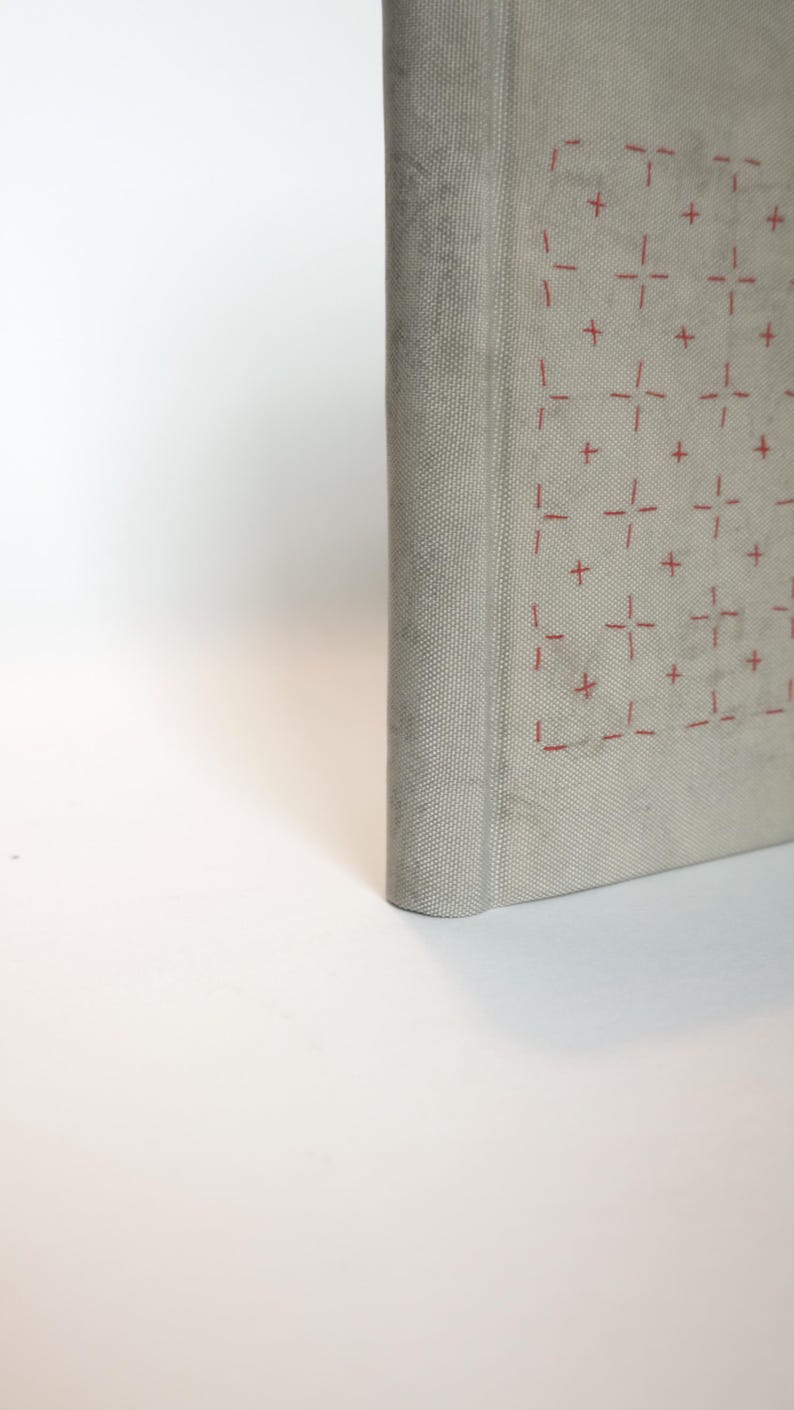 May include: A light gray book with a textured cover. The cover features a repeating pattern of red cross-like designs. The spine is rounded, and the book appears to be closed, with the pages unseen.