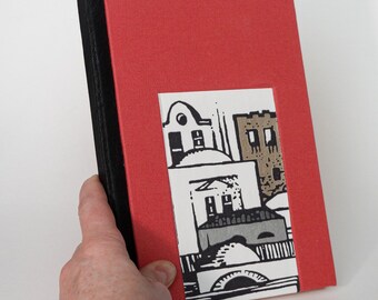 May include: A red hardcover journal with a black spine and a purple ribbon bookmark. The front cover features a black and white illustration of a cityscape with buildings and a bridge.