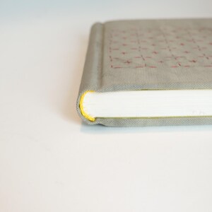 May include: A closed, rectangular book with a light gray cover featuring a grid pattern of red stitching. The edges of the pages are white, and the spine is a darker gray. The book's edges are highlighted with yellow.