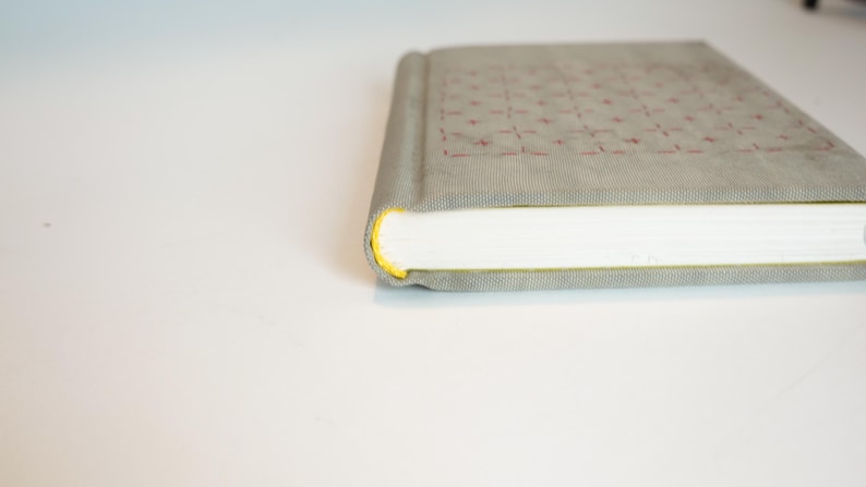 May include: A closed, rectangular book with a light gray cover featuring a grid pattern of red stitching. The edges of the pages are white, and the spine is a darker gray. The book's edges are highlighted with yellow.