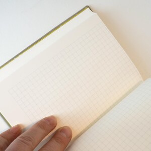 May include: Open notebook with grid paper. The notebook has a green spine and a cream-colored cover. The pages are open, revealing the grid pattern. A hand is visible, holding the notebook open on a white surface.