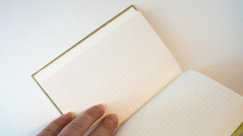 May include: Open notebook with grid paper. The notebook has a green spine and a cream-colored cover. The pages are open, revealing the grid pattern. A hand is visible, holding the notebook open on a white surface.