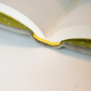 May include: An open notebook with graph paper pages. The cover is gray fabric with a yellow spine and a green polka dot interior lining. The notebook is on a white surface.