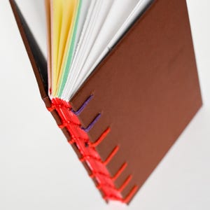 Op de afbeelding: Een handgemaakt notitieboek met een bruine kaft en kleurrijke pagina's. De binding is gestikt met rood en paars garen. De pagina's zijn een mix van wit, geel, groen en roze.
