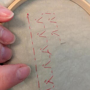 May include: Embroidery hoop holding light green fabric with red stitching. The stitching forms the letter "B" with a dotted line and arrow-like shapes. The wooden hoop is partially visible.