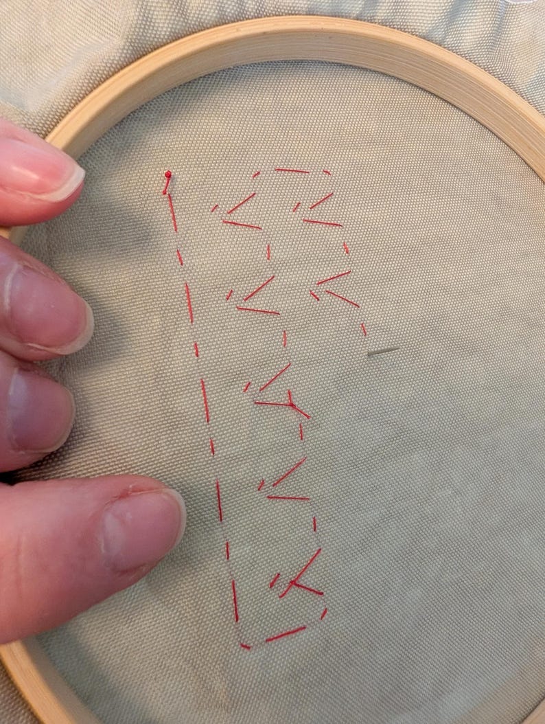 May include: Embroidery hoop holding light green fabric with red stitching. The stitching forms the letter "B" with a dotted line and arrow-like shapes. The wooden hoop is partially visible.