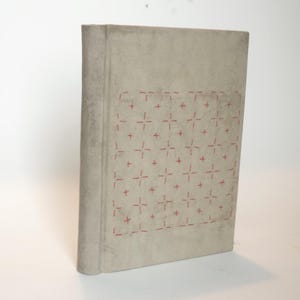 Könnte beinhalten: Ein Hardcover-Buch mit hellgrauem Einband. Der Einband zeigt ein Gittermuster aus roter Kreuzstichstickerei. Der Buchrücken ist dunkler grau. Das Buch steht aufrecht vor einem weißen Hintergrund.