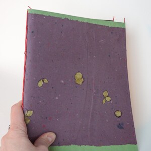 May include: A handmade journal with a purple cover speckled with flecks of color and embedded with dried leaves. The journal is bound with red thread and has a green border along the top and bottom edges.