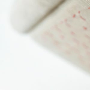 May include: Close-up of a beige fabric with red stitching in a geometric pattern. The stitching creates a series of small, connected shapes. The fabric appears to be soft and textured, with a slight sheen.