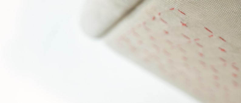 May include: Close-up of a beige fabric with red stitching in a geometric pattern. The stitching creates a series of small, connected shapes. The fabric appears to be soft and textured, with a slight sheen.