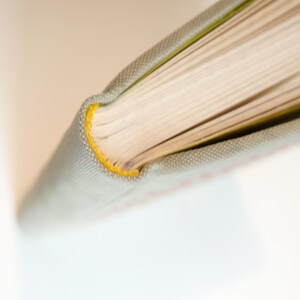 May include: Close-up of a book's spine and pages. The book has a light gray cover with a yellow stitched edge. The pages are cream-colored, and the book is open, revealing the paper's texture.