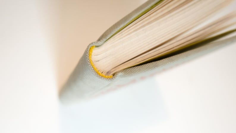 May include: Close-up of a book's spine and pages. The book has a light gray cover with a yellow stitched edge. The pages are cream-colored, and the book is open, revealing the paper's texture.