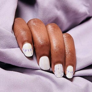 May include: Close-up of a hand with painted nails. The nails are painted white with a design of purple and blue geometric shapes and silver stars. The hand rests on a lavender fabric.