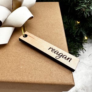 Puede incluir: Una caja de regalo marrón con un lazo blanco y una etiqueta de regalo de madera con el nombre "reagan" grabado en ella.