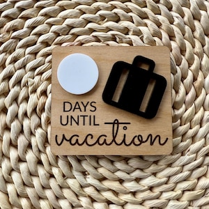 May include: A wooden countdown block with the text "DAYS UNTIL vacation". It features a white circle and a black suitcase cutout. The block is set against a woven background.
