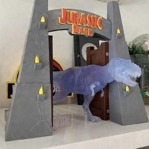 May include: A Jurassic Park themed playset featuring a T-Rex figure emerging from the iconic park entrance. The entrance is gray with yellow flame accents and the Jurassic Park logo. A toy car and Christmas card are in the background.