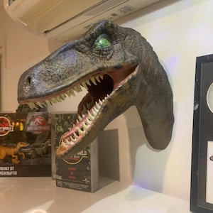 Könnte beinhalten: Eine an der Wand montierte Velociraptor-Dinosaurierkopf-Skulptur mit strukturierter brauner und grüner Haut, einem weit geöffneten Maul mit scharfen weißen Zähnen und einem sichtbaren grünen Auge. Die Skulptur befindet sich neben Jurassic Park-Sammlerstücken.
