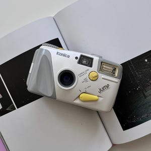 May include: A white and grey Konica Jump weatherproof 35mm film camera with a 35mm F4 lens. The camera features yellow accents on the shutter button and flash switch, with text reading Konica, Konica Lens 35mm F4, and Jump Weatherproof printed on the front.