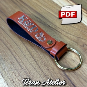 May include: A brown leather keychain with a gold ring and the text "K&D 23.11.22" engraved in silver. The keychain has a black leather strap and a silver ring with a design of two intertwined rings.
