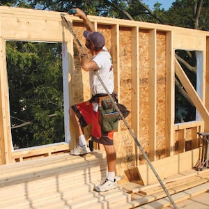 May include: A construction worker uses a long tool to secure a window frame in a wooden wall. The wall is made of plywood and has a rough texture. The window frame is made of white painted wood. The worker is wearing a blue shirt, red shorts, and a tool belt.