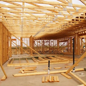 Op de afbeelding: Een houten framehuis in aanbouw. Het interieur van het huis is zichtbaar, met de houten balken en steunen. Het huis is in de vroege stadia van de bouw, zonder muren of dak.