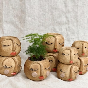 May include: A set of seven ceramic planters shaped like faces with closed eyes and red lips. The planters are stacked in pairs and feature a variety of facial details, including eyelashes, noses, and cheeks. One planter has a small green plant growing inside.