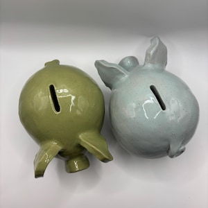 Può includere: Due salvadanai in ceramica, uno verde oliva maculato e l'altro azzurro maculato. Ogni salvadanaio presenta un corpo arrotondato, un muso, due orecchie e una stretta fessura per le monete sulla parte superiore. Le superfici hanno una finitura lucida.