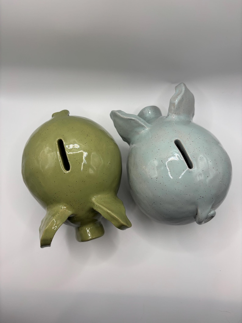 Puede incluir: Dos huchas de cer&aacute;mica, una en verde oliva moteado y la otra en azul claro moteado. Cada hucha presenta un cuerpo redondeado, un hocico, dos orejas y una ranura estrecha para monedas en la parte superior. Las superficies tienen un acabado brillante.