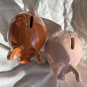 Puede incluir: Dos huchas de cer&aacute;mica, una en color marr&oacute;n terracota oscuro brillante y la otra en rosa pastel brillante, ambas con acabado moteado, forma redondeada, peque&ntilde;as orejas que sobresalen y una ranura vertical para monedas en la parte superior.