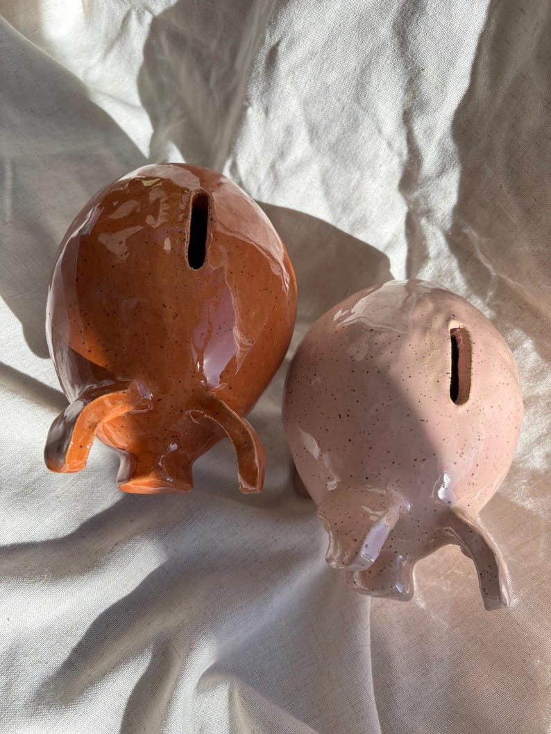 Puede incluir: Dos huchas de cer&aacute;mica, una en color marr&oacute;n terracota oscuro brillante y la otra en rosa pastel brillante, ambas con acabado moteado, forma redondeada, peque&ntilde;as orejas que sobresalen y una ranura vertical para monedas en la parte superior.