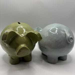 Puede incluir: Dos huchas de cer&aacute;mica con un acabado brillante y moteado. Una es de color verde oliva y la otra es de color azul claro. La hucha azul tiene una peque&ntilde;a cola rizada y la palabra KADE estampada en letras negras en el lateral. Ambas tienen ranuras.