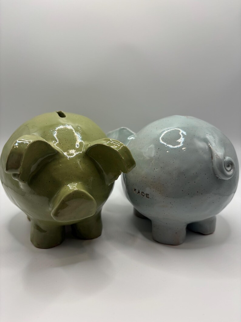 Puede incluir: Dos huchas de cer&aacute;mica con un acabado brillante y moteado. Una es de color verde oliva y la otra es de color azul claro. La hucha azul tiene una peque&ntilde;a cola rizada y la palabra KADE estampada en letras negras en el lateral. Ambas tienen ranuras.