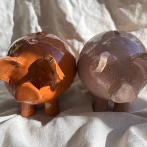 Puede incluir: Dos huchas de cer&aacute;mica, una en color naranja quemado brillante y la otra en rosa pastel suave, ambas con un acabado moteado, cuerpos redondeados, orejas peque&ntilde;as y una ranura para monedas en la parte superior, colocadas una al lado de la otra.