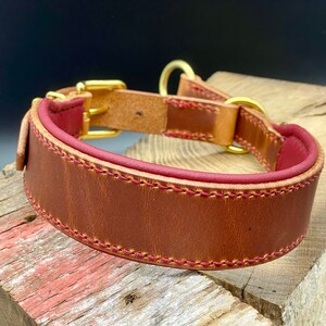 Könnte beinhalten: Ein braunes Lederhundekragen mit rotem Lederfutter und goldenen Metallbeschlägen. Das Halsband ist mit einem kontrastierenden Faden vernäht.