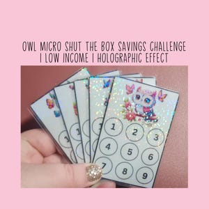 Può includere: Una mano tiene in mano diverse carte bianche con effetto olografico. Ogni carta presenta un'illustrazione di gufo, farfalle e i numeri 1-9. Il testo sulle carte recita "OWL MICRO SHUT THE BOX SAVINGS CHALLENGE I LOW INCOME I HOLOGRAPHIC EFFECT."