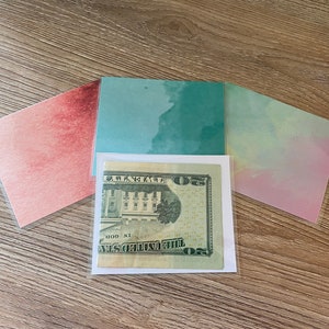 May include: A $50 US bill is displayed in a clear plastic sleeve. The bill is surrounded by three colorful sheets of paper with watercolor-like designs. The sheets are pink, blue, and a light pink.