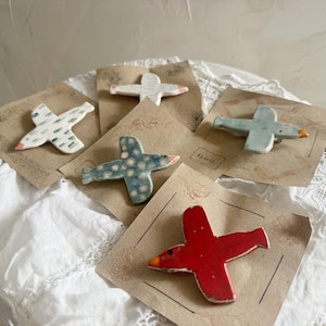 Puede incluir: Cinco broches de cerámica con forma de pájaro presentados en tarjetas de papel marrón. Los broches tienen varios diseños, incluyendo un pájaro rojo sólido con pico naranja, un pájaro azul con puntos blancos, uno azul claro, uno blanco y uno blanco con rayas verdes.
