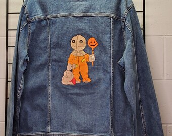 May include: A blue denim jacket with an embroidered patch of a character holding a candy corn lollipop. The character is wearing an orange jumpsuit and has a stitched-up face.