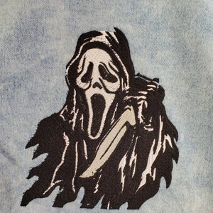 May include: Black and white embroidered patch of a ghost figure wearing a hooded robe and holding a knife. The ghost has a white mask with a wide open mouth and a scream.