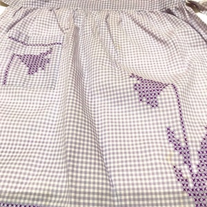 Op de afbeelding: Een paarse en witte gingham schort met paarse geborduurde bloemen. Het schort heeft een zak aan de linkerkant.