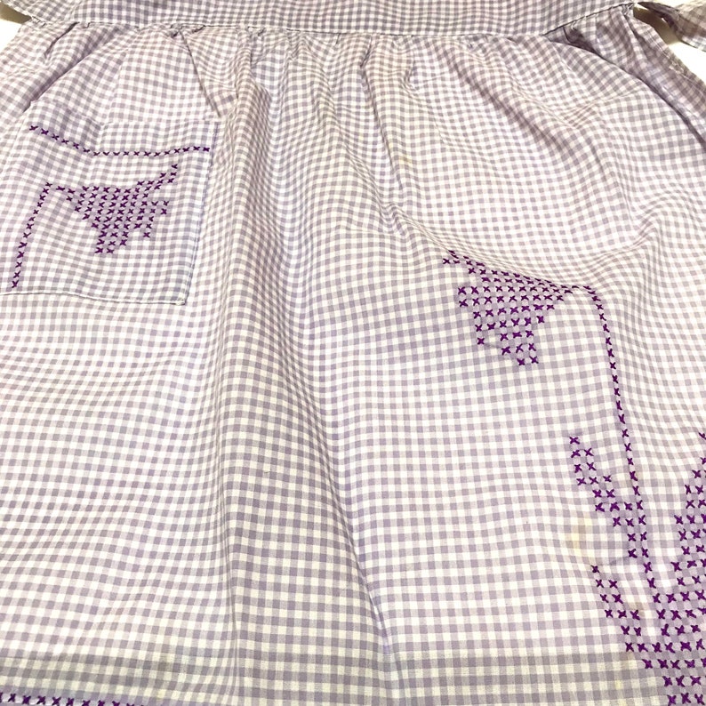 Op de afbeelding: Een paarse en witte gingham schort met paarse geborduurde bloemen. Het schort heeft een zak aan de linkerkant.