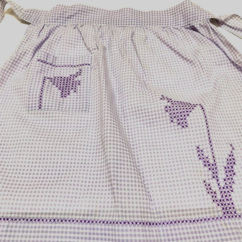 Op de afbeelding: Een witte gingham schort met paarse geborduurde bloemen en een zak. Het schort heeft een paarse gingham zoom.
