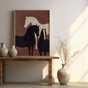 K&ouml;nnte beinhalten: Gerahmter Kunstdruck mit drei stilisierten Pferden in Schwarz und Creme vor einem rotbraunen Hintergrund. Das Kunstwerk wird &uuml;ber einem Holztisch mit zwei beigen Vasen pr&auml;sentiert. Eine Vase mit getrockneten Zweigen steht daneben.