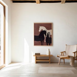 Puede incluir: Una impresión artística enmarcada con tres caballos en blanco y negro sobre un fondo granate. La obra de arte cuelga sobre un banco de madera con un jarrón blanco. A la derecha hay una silla de madera con una manta crema.