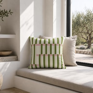 May include: A square throw pillow featuring a vertical striped pattern in shades of olive green, cream, and pale pink. The design consists of alternating green and cream stripes with horizontal pink accents intersecting the vertical lines.