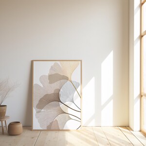 May include: A framed art print featuring a botanical design with overlapping, stylized ginkgo leaves in shades of beige, tan, and white. The artwork is displayed against a white wall, with a wooden floor and a large window providing natural light.