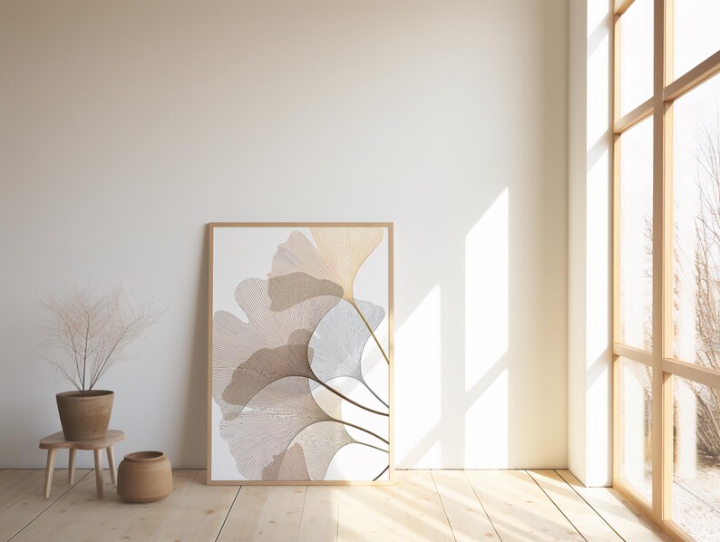 May include: A framed art print featuring a botanical design with overlapping, stylized ginkgo leaves in shades of beige, tan, and white. The artwork is displayed against a white wall, with a wooden floor and a large window providing natural light.