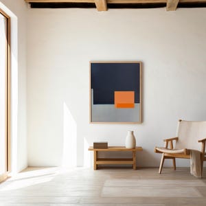 May include: An interior scene with a large framed abstract painting in navy, grey, and orange. A wooden bench holds a white vase and books. A wooden chair with a cream seat and a blanket is to the right. The room has a minimalist design.