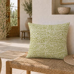 May include: A square throw pillow featuring a white base with a repeating pattern of small, olive green dots arranged in a grid. The pillow has a soft, fabric texture and is positioned on a woven bench next to a straw hat in a bright, sunlit space.